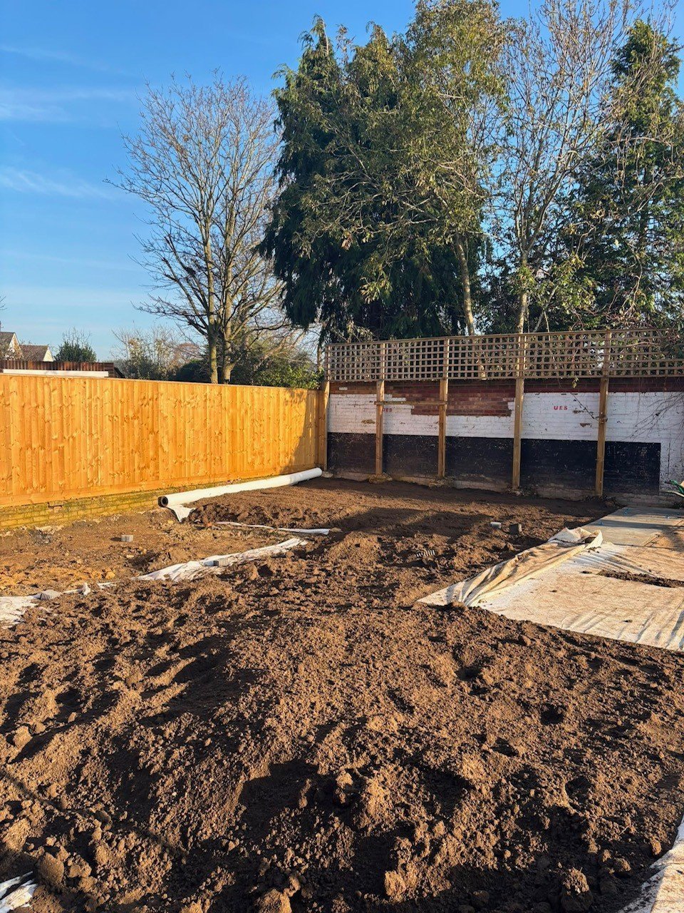 Backyard garden plot with soil, orange fence, wooden structures, and bare trees under blue sky