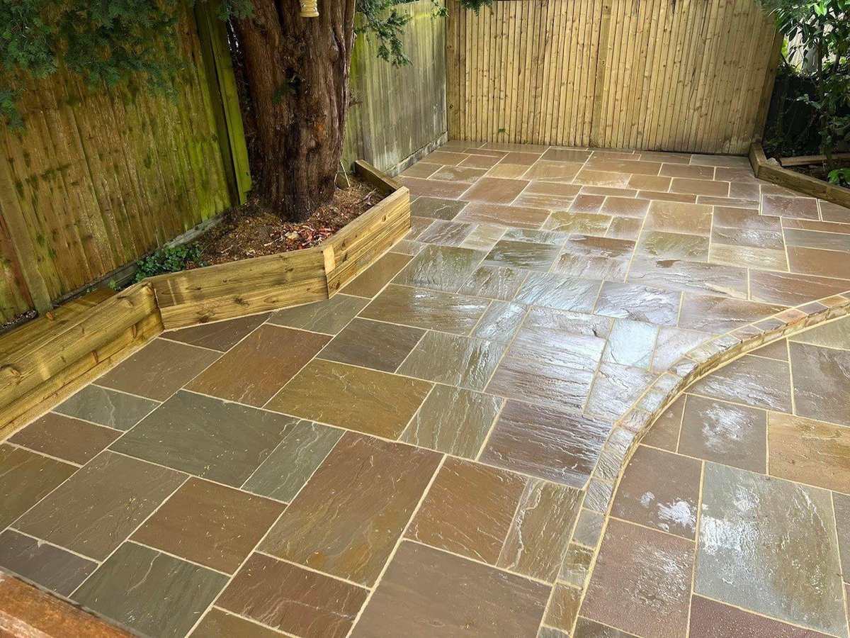 Newly installed patio with tan and brown flagstone tiles in a herringbone pattern, bordered by wooden edging and fencing