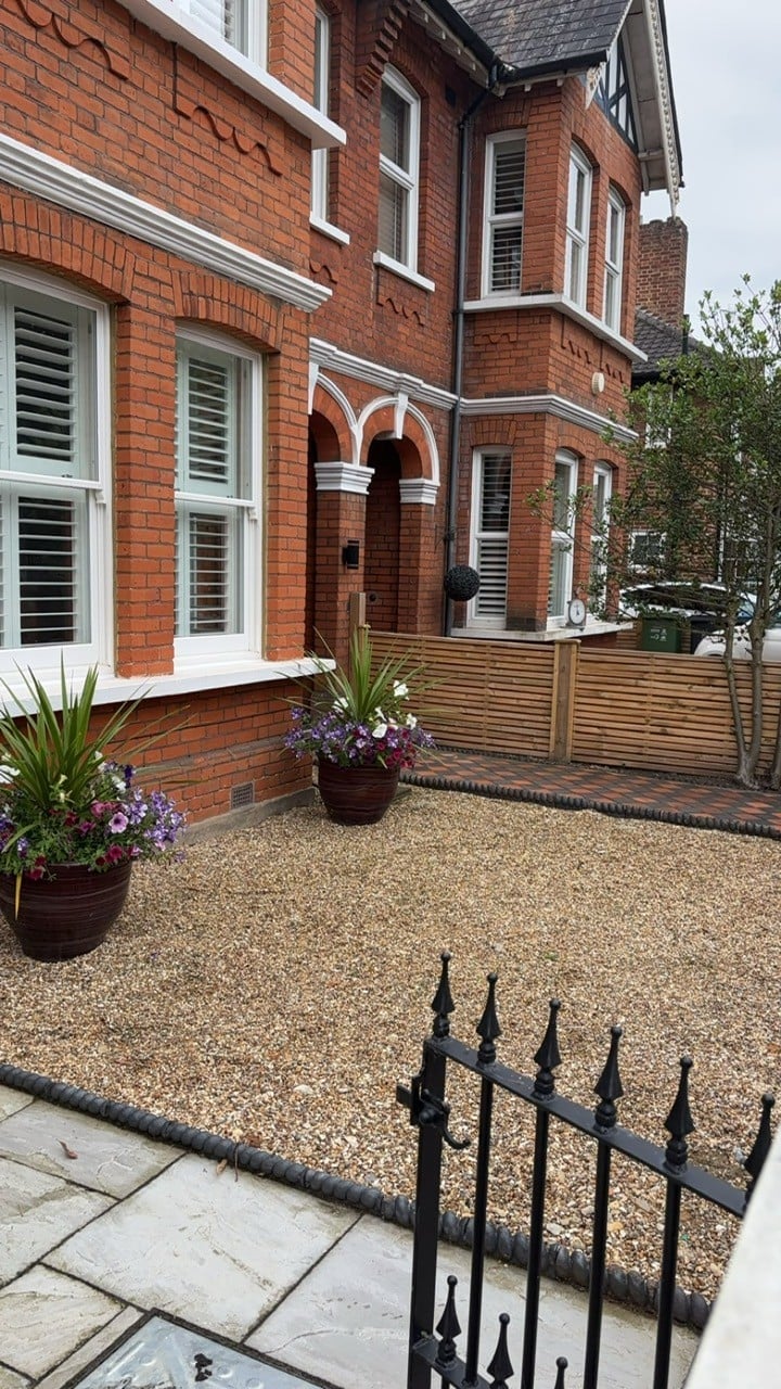 Red brick Victorian house with gravel courtyard, potted flowers, wooden fence, and black iron fence in foreground