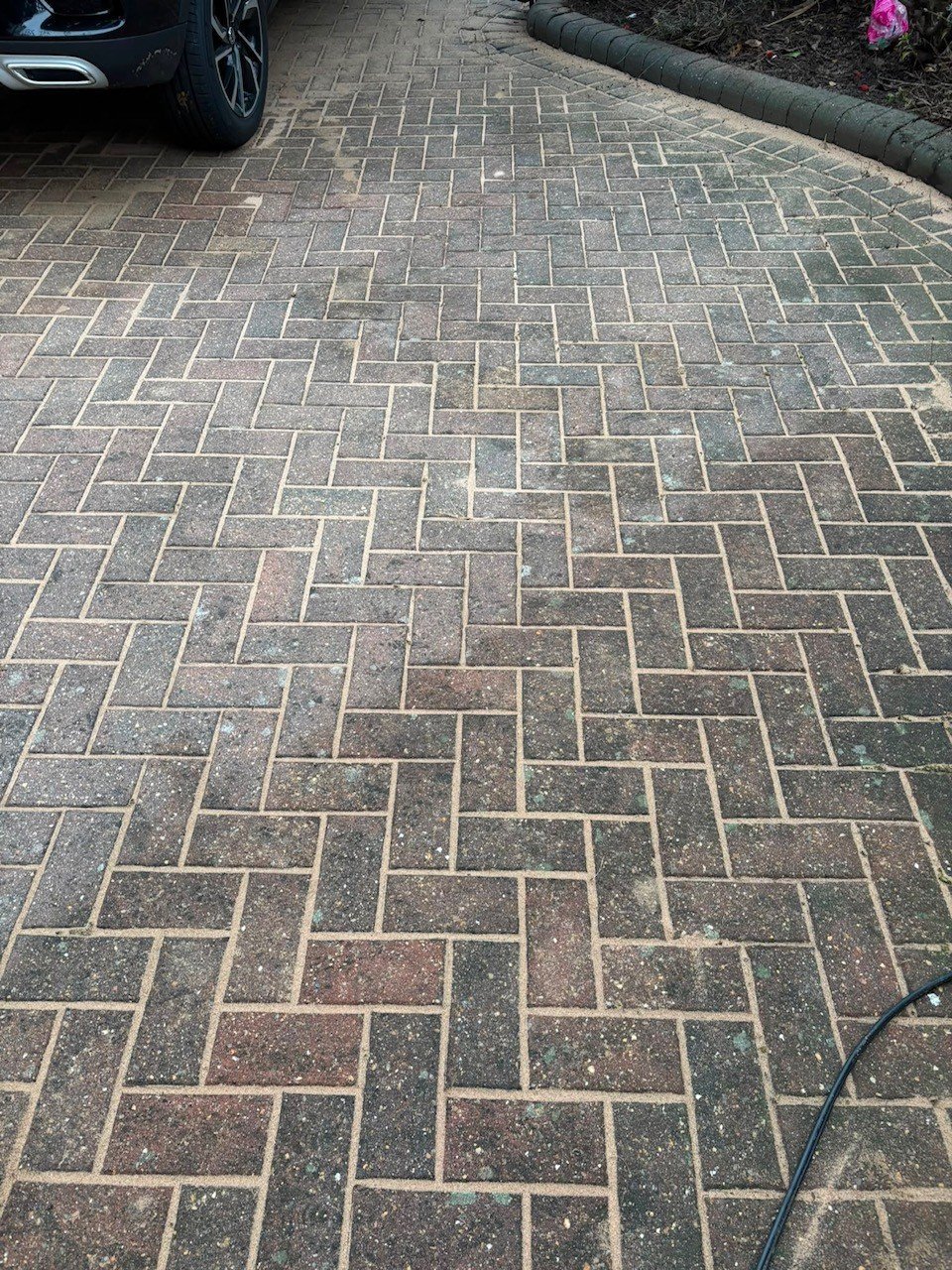 Herringbone patterned brick driveway with moss growth, parked car visible at top left, pink flowers and log at edges
